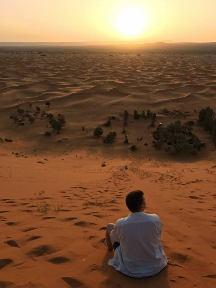Photograph of a man in a white shirt sitting in orange colored sand looking at the sun.